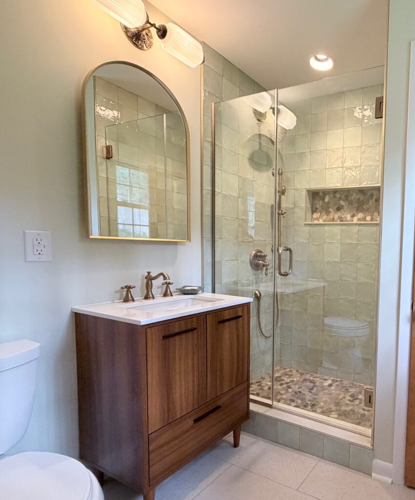 Primary bathroom remodel in Raleigh with soft green shower tile, pebble stone shower floor, frameless glass enclosure, and warm wood vanity.